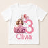 A white t-shirt with a cartoon image of a girl in a pink dress and a crown, along with balloons and the number 3. The text below the image says "Olivia".