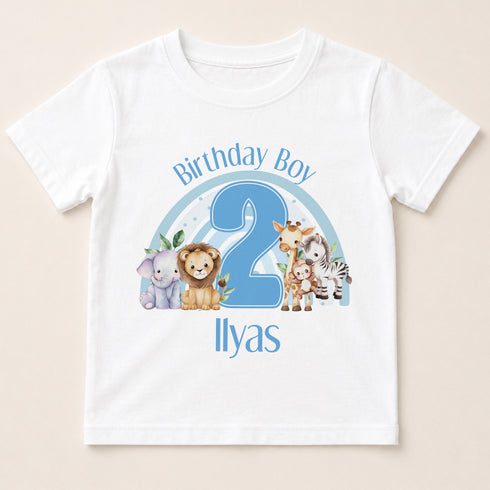 A white t-shirt with a graphic design featuring a blue number 2 surrounded by various cartoon animal characters, including a lion, zebra, giraffe, and elephant. The text "Birthday Boy 2" is written above the number, and "Ilyas" is written below the number.