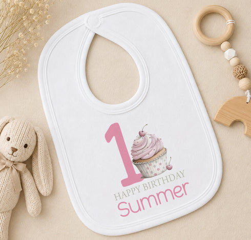 Personalised first birthday bib with pink cupcake design and custom name, styled with baby toys