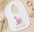 Personalised first birthday bib with pink cupcake design and custom name, styled with baby toys
