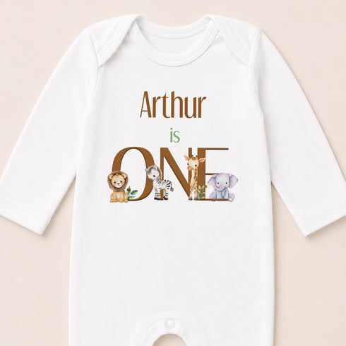 A white onesie with the text "Arthur is ONE" and illustrations of a lion, zebra, and elephant.