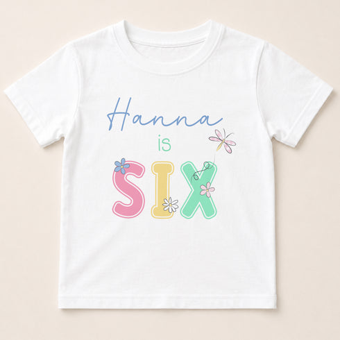 A white t-shirt with the text "Hannah is Six" printed on it in a colorful, stylized font.