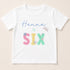 A white t-shirt with the text "Hannah is Six" printed on it in a colorful, stylized font.