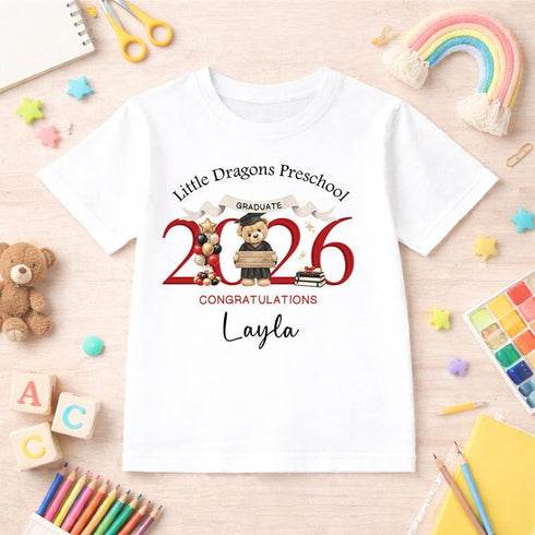 White children’s T-shirt featuring a personalised red 2026 teddy bear preschool graduation design, photographed with colourful nursery and craft accessories.