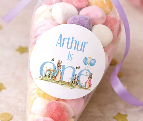 A clear plastic container filled with colorful candies, with a round label on it that says "Arthur is one" and features illustrations of rabbits and balloons.