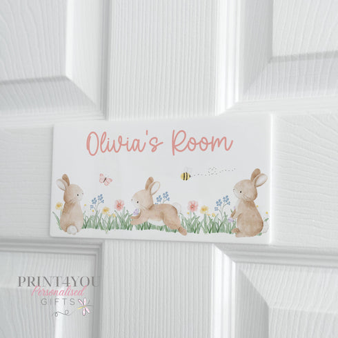 White door with a rectangular sign reading 'Olivia's Room' in pink cursive font. The sign features three brown bunnies, flowers, a butterfly, and a bee in a grassy field.