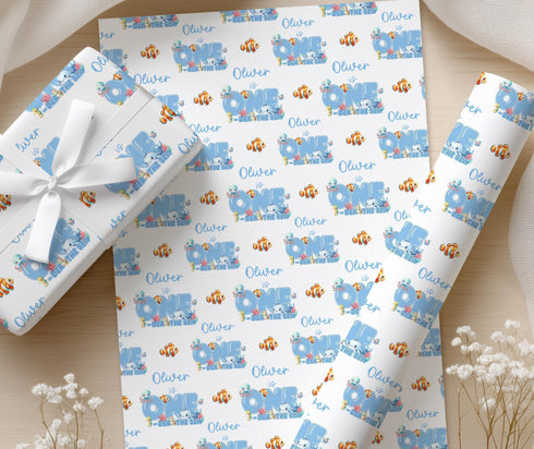 a set of wrapping paper with a repeating pattern of the name "Oliver" and illustrations of fish, including Nemo, against a blue background. The wrapping paper is displayed on a wooden surface, with some white flowers visible in the background.