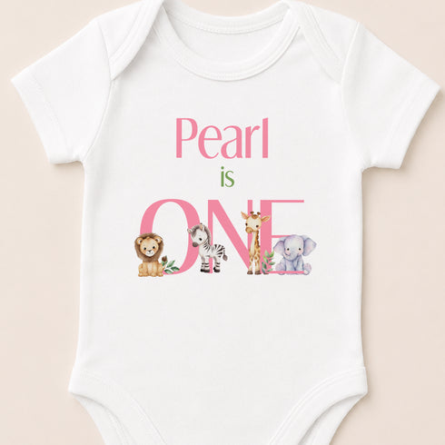 A white baby onesie with the text "Pearl is ONE" printed on it, featuring illustrations of a lion, zebra, giraffe, and elephant.