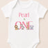 A white baby onesie with the text "Pearl is ONE" printed on it, featuring illustrations of a lion, zebra, giraffe, and elephant.