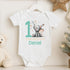 Personalised 1st birthday baby outfit with cute elephant, party hat, stars, teal number 1 and custom name, shown on bib, vest, sleepsuit, t-shirt and white pyjamas.