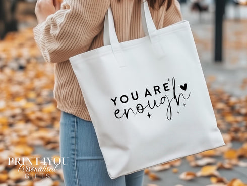 A white cotton tote bag which says "you are enough" in black text. The bag is being shown over a ladies shoulder on an autumn day with leaves on the ground.