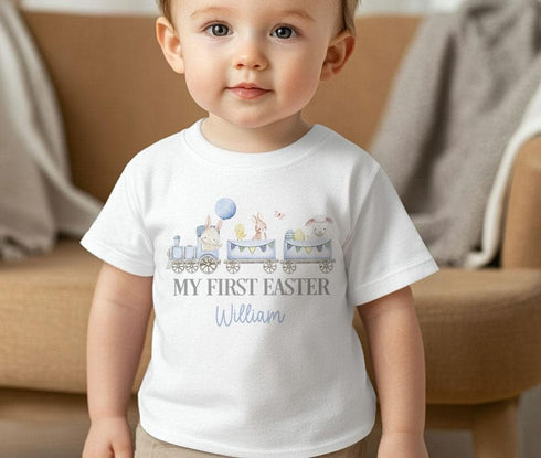 Image of a toddler wearing a personalized white t-shirt with a festive Easter design. The design features a train with three carriages, each carrying Easter-themed characters: a bunny with a blue balloon, a bunny and a chick with Easter eggs, and a lamb. Above the train, the text 'MY FIRST EASTER' is printed in large grey letters, and the name 'William' appears below in blue cursive. The background includes a blurred indoor setting, suggesting a casual and cosy environment