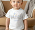 Image of a toddler wearing a personalized white t-shirt with a festive Easter design. The design features a train with three carriages, each carrying Easter-themed characters: a bunny with a blue balloon, a bunny and a chick with Easter eggs, and a lamb. Above the train, the text 'MY FIRST EASTER' is printed in large grey letters, and the name 'William' appears below in blue cursive. The background includes a blurred indoor setting, suggesting a casual and cosy environment