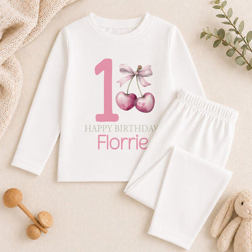 Personalised 1st birthday girl outfit with pink number 1, cute cherry bow design, happy birthday text and custom name, shown on bib, vest, sleepsuit, t-shirt and white pyjamas.