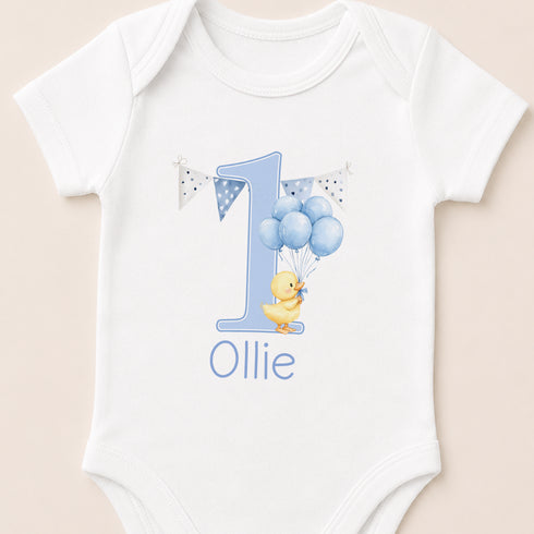 A white onesie with a blue number one and a yellow duck, along with the text "Ollie" printed on it.