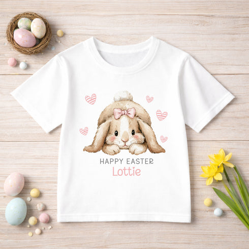 A white t-shirt with a bunny graphic and the text "HAPPY EASTER LOTTIE" printed on it, along with a bow and hearts. The t-shirt is displayed on a wooden surface, surrounded by Easter decorations such as eggs, flowers, and other Easter-themed items.