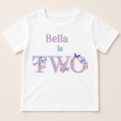 A white t-shirt with the text "Bella is two" printed on it, along with a design of butterflies.