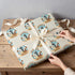 A person's hands are wrapping a gift box with a patterned wrapping paper featuring the word "RODEO" and a cowboy silhouette.