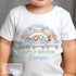 Child's white t-shirt featuring a colorful train with animals (zebra, lion, elephant, monkey, giraffe) and a rainbow with bunting flags above. Text reads: 'HAPPY 1st BIRTHDAY Roman' in green and blue.