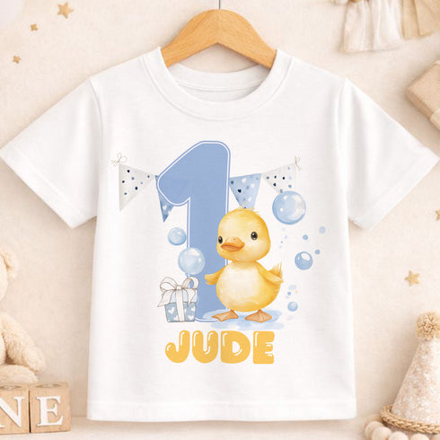 A white t-shirt with a cartoon image of a yellow duck and the number "1" printed on it, along with the text "JUDE".