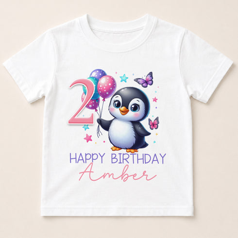 A white t-shirt with a cartoon penguin holding balloons and surrounded by butterflies, along with the text "2 HAPPY BIRTHDAY AMBER".
