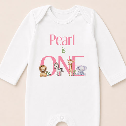 A white baby onesie with the text "Pearl is one" and illustrations of various animals, including a lion, zebra, giraffe, and elephant.