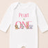 A white baby onesie with the text "Pearl is one" and illustrations of various animals, including a lion, zebra, giraffe, and elephant.