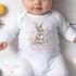 Image of a baby wearing a white onesie with an Easter-themed design. The onesie features a charming illustration of a bunny surrounded by colourful flowers. Below the illustration, the text reads 'MY FIRST EASTER' in grey and 'Savannah' in pink cursive with a small purple heart beneath the name.