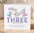 A birthday card with illustrations of three dinosaurs and balloons, along with the text "THREE" and "Happy third birthday to a roaring son".