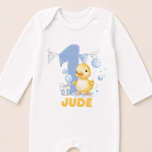 A white onesie with a cartoon image of a yellow duck and the text "JUDE" printed on it.