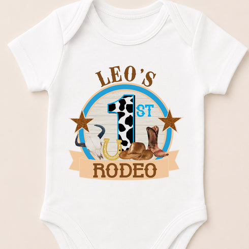 A white baby onesie with a graphic design featuring a cowboy hat, a horseshoe, and the text "LEO'S 1ST RODEO" printed on it.