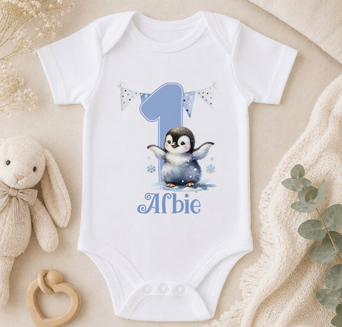 A white onesie with a penguin design and the number 1, along with the text "Afbie" printed on it, is displayed on a bed. There is also a stuffed animal and some greenery in the background.
