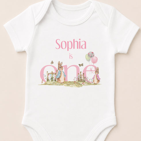 A white baby onesie with a personalized text and illustrations related to a first birthday celebration.