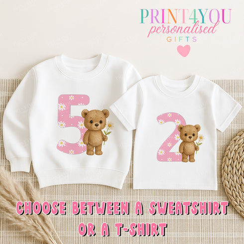 two white t-shirts with a pink number 5 and pink number 2, each featuring a teddy bear holding a flower. The t-shirts are placed on a beige background, and there is text that reads "Choose between a sweatshirt or a T-shirt".