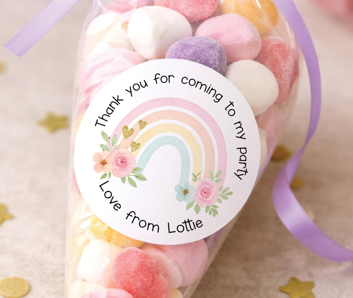 Round sticker printed with a pastel rainbow, watercolour flowers and gold hearts, reading Thank you for coming to my party, Love from Lottie, stuck on a bag of pastel party sweets tied with a lilac ribbon.