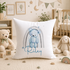 A white pillow with a blue bear and a rainbow design, surrounded by stuffed animals and toys in a nursery setting.