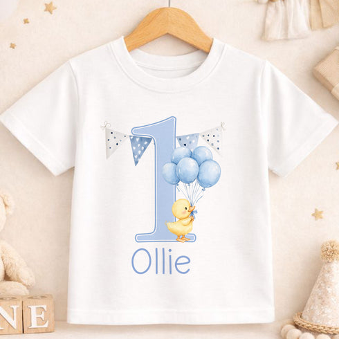 A white t-shirt with a blue number one and a yellow duck holding blue balloons, along with the name "Ollie" printed on it.