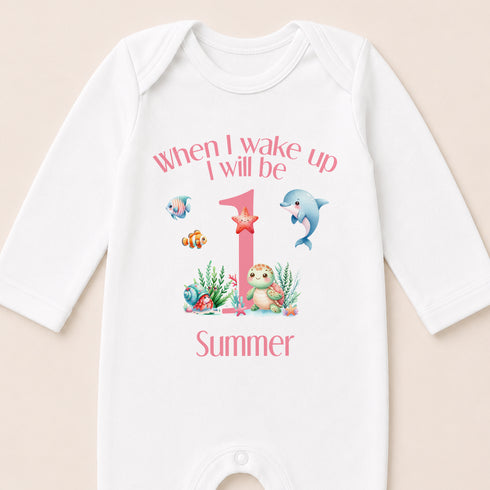 A white onesie with a printed design on it, featuring a pink number 1, a blue dolphin, and various marine life. The text on the onesie says "When I wake up I will be 1 Summer".