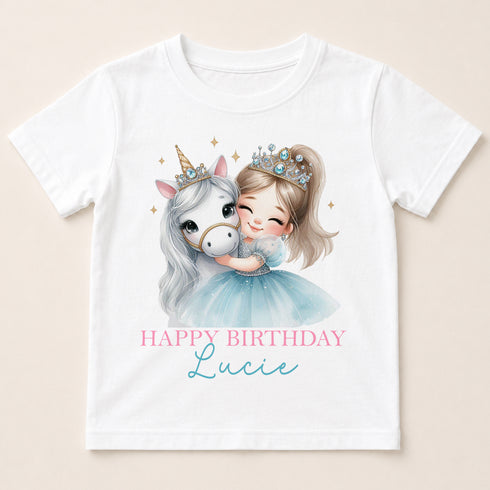 A white t-shirt with a printed design featuring a young girl wearing a crown and hugging a white unicorn. The text below the image reads "Happy Birthday Lucie".