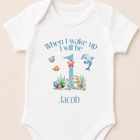 A white onesie with a personalized design for a baby's first birthday. The design includes a blue number 1, a dolphin, a starfish, and other marine life. The text above the design says "When I wake up I will be Jacob".
