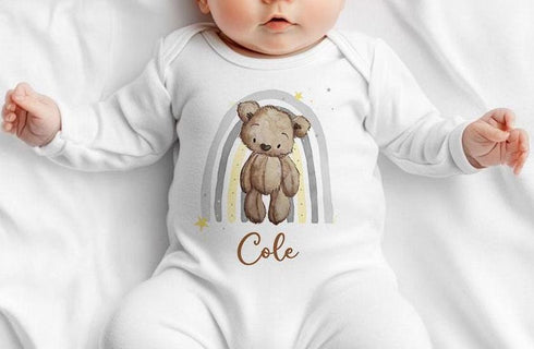 Alt text: A baby is lying on a white blanket, wearing a white onesie with a design featuring a cute brown teddy bear and a rainbow with stars. The name "Cole" is written in brown cursive text below the teddy bear.
