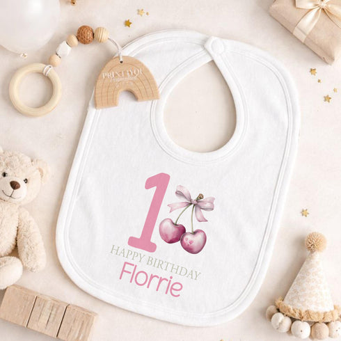 Personalised 1st birthday girl outfit with pink number 1, cute cherry bow design, happy birthday text and custom name, shown on bib, vest, sleepsuit, t-shirt and white pyjamas.