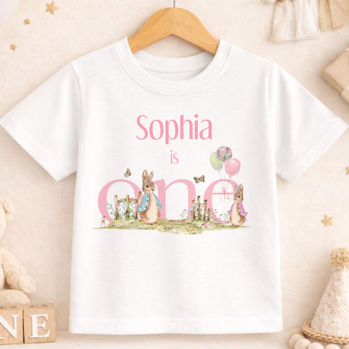A white t-shirt with a personalized text and illustrations, including a rabbit and balloons, is displayed on a wooden hanger.