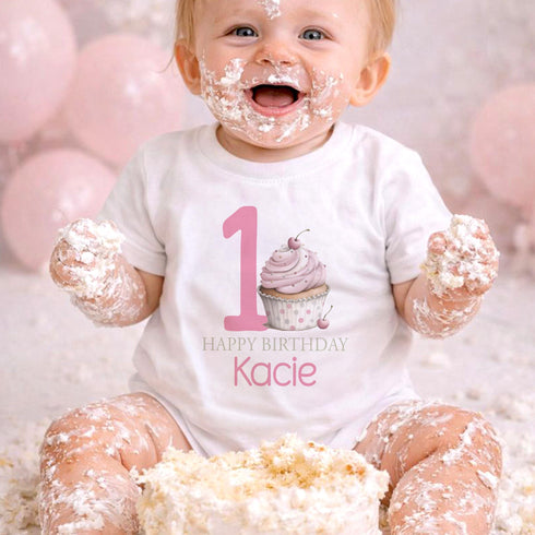 Personalised 1st birthday girl outfit with pink number 1, cute cupcake design, happy birthday text and custom name, shown on sleepsuit, t-shirt, vest, pyjamas and bib.