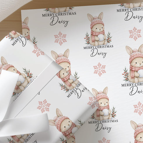 Personalised Christmas wrapping paper featuring a cute bunny rabbit in a pink knitted hat and scarf, custom name Christmas gift wrap with festive snowflakes.
