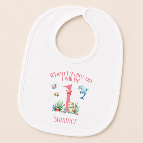 A white baby bib with a playful design featuring a dolphin, fish, and other marine life, along with the text "When I wake up I will be 1 Summer".