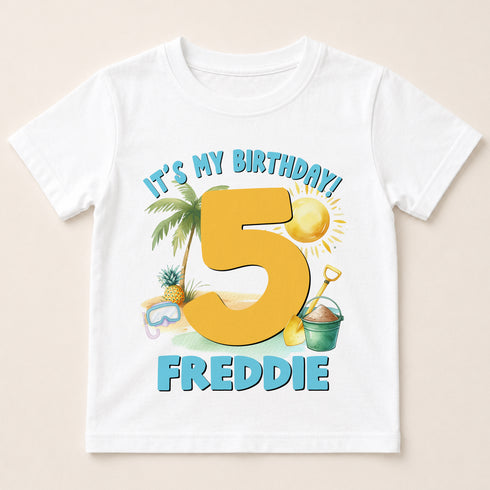 A white t-shirt with a graphic design featuring a large number 5, a palm tree, a beach scene, and a bucket. The text above the number reads "It's my birthday!" and below the number is the name "FREDDIE".