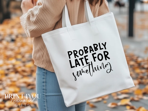 A white cotton tote bag which says "Probably late for something" in black text. The bag is being shown over a ladies shoulder on an autumn day with leaves on the ground.