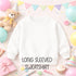 A white long-sleeved sweatshirt is displayed among a variety of colorful party decorations, including balloons, a teddy bear, and a cupcake.