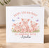 A birthday card with a group of five cute pink pigs sitting together in a grassy field, surrounded by flowers and butterflies. The card has a personalized message that reads "Happy 6th Birthday to a wonderful sister Amelia".